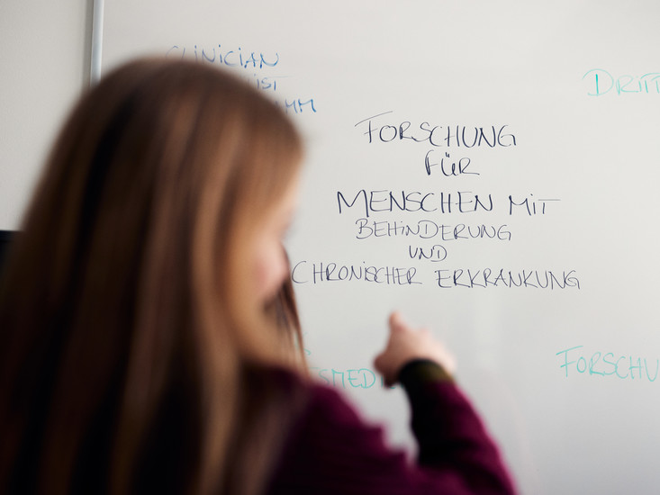 Overshoulder Shot: Eine Forschende zeigt auf ein Whiteboard, auf dem der Text "Forschung für Menschen mit Behinderung und chronischer Erkrankung" geschrieben steht.