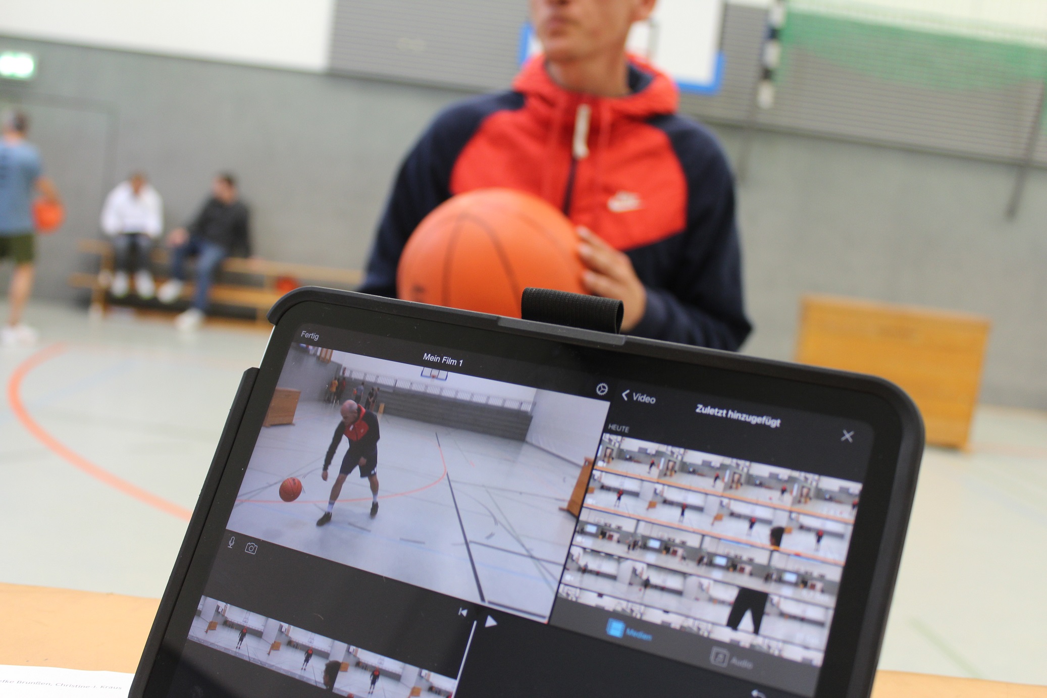 Auf einem Bildschirm werden Videoaufnahmen aus der Sporthalle gezeigt, hinter dem Bildschirm steht ein Mann mit einem Basketball in den Händen