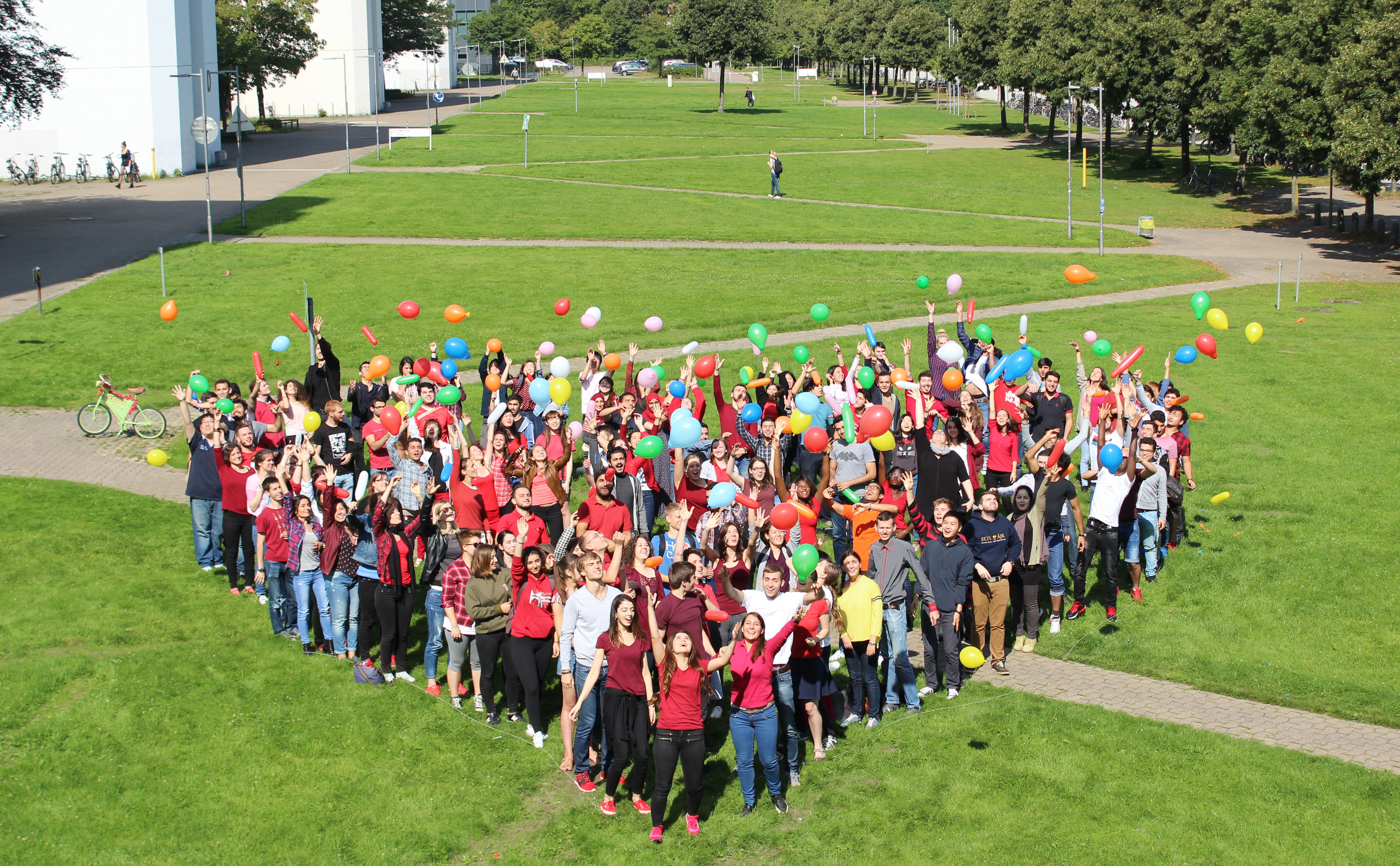 Teilnehmende der Sommerkurse stehen in Herzform zusammen und werfen Luftballons in die Luft