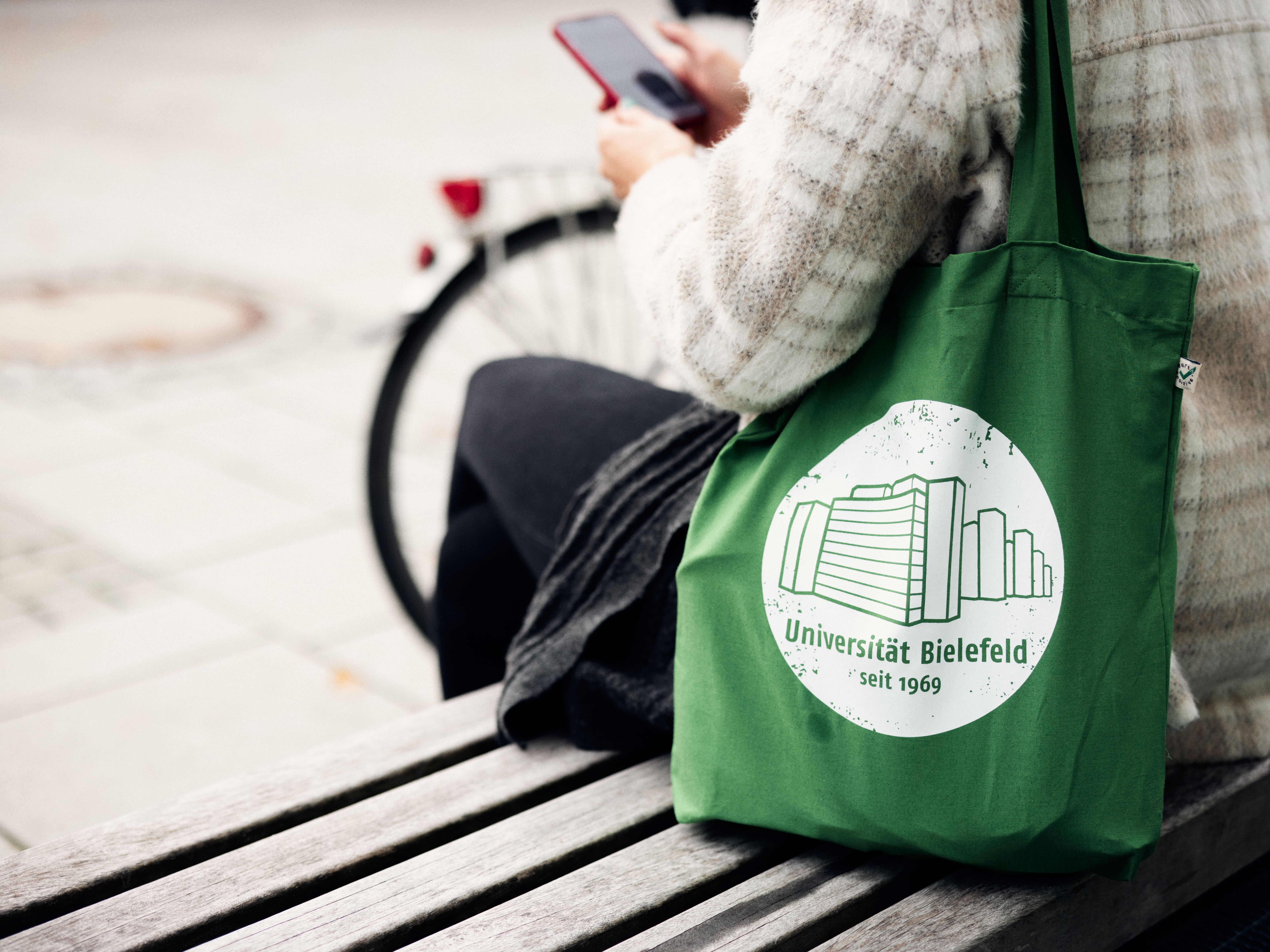 Person sitzt auf einer Bank, schaut auf ihr Handy und hat einen grünem Stoffbeutel und Logo der Universität an der Schulter hängen.