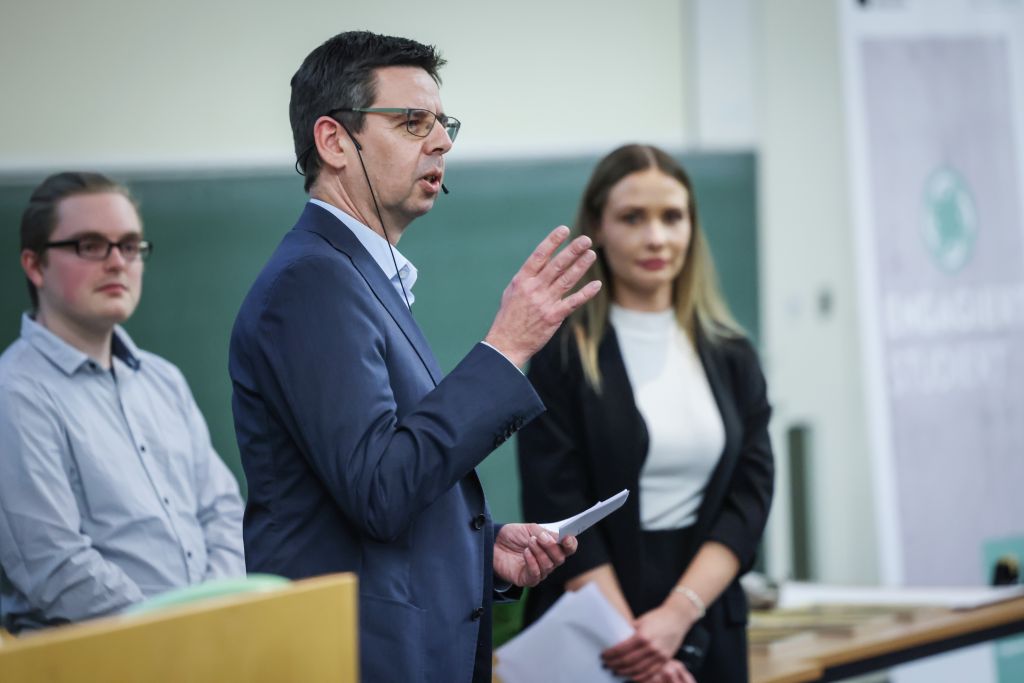 Dr. André Fleer hält eine Rede, die Moderator*innen stehen im Hintergrund.