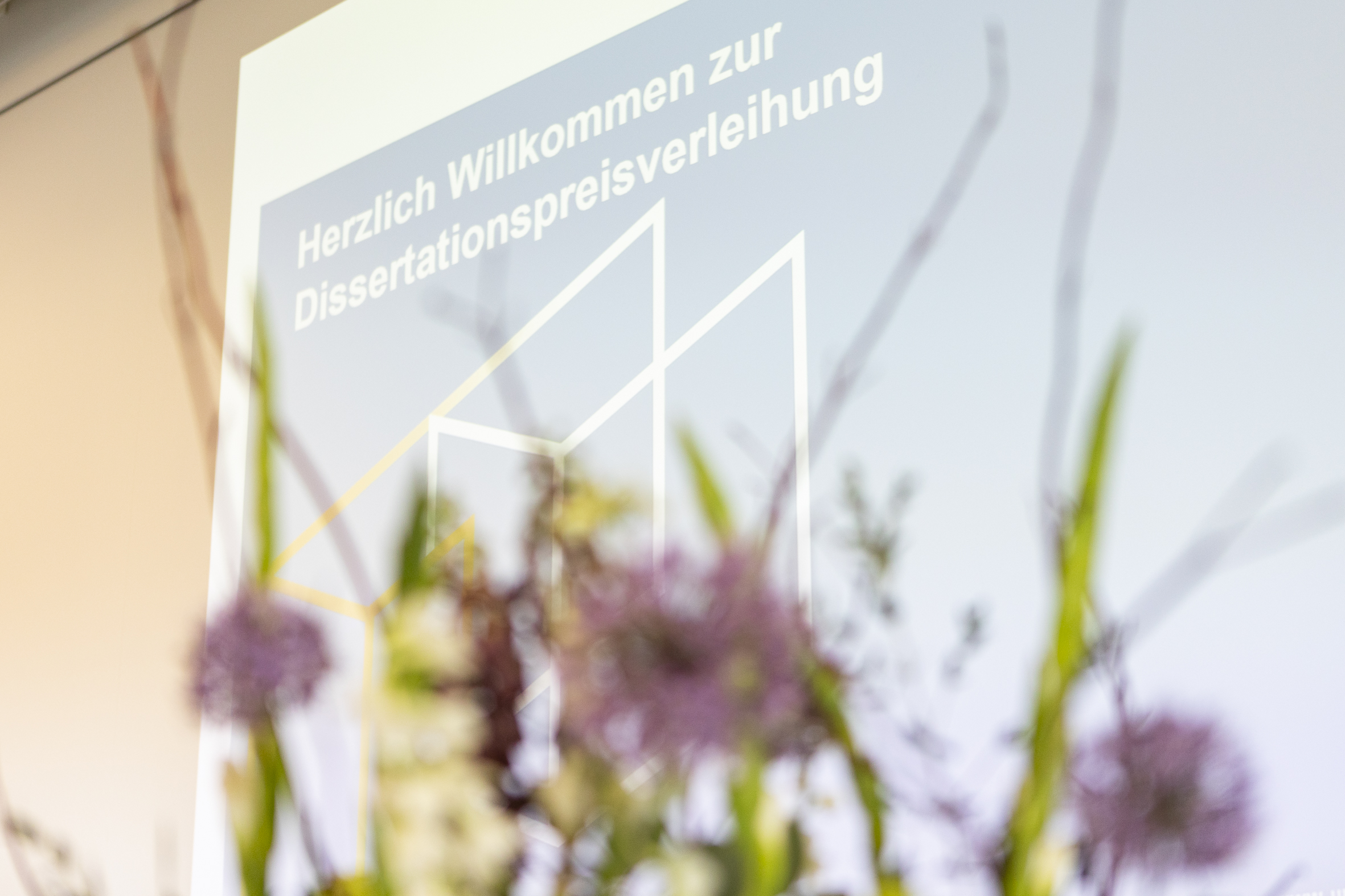 Zu sehen ist ein Plakat des Dissertationspreises mit einem Blumenarrangement im Vordergrund.