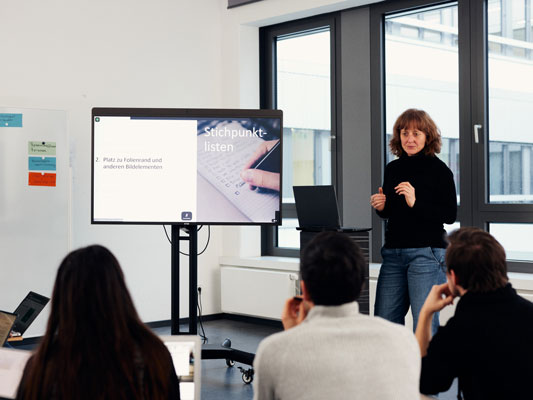 In einer Workshopsituation erklärt eine Frau einer Gruppe von Wissenschaftler*innen etwas