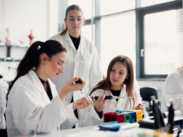 Schülerinnen füllen Laborproben mit Pipetten um, hinter ihnen setht eine Studentin