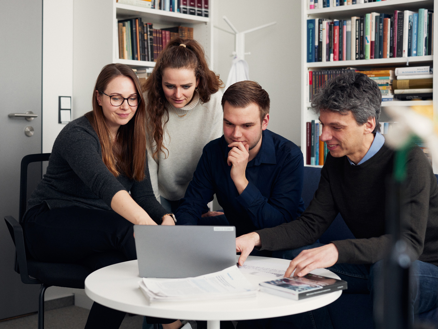 Eine Gruppe junger Forschender sitzt vor Bücherregalen um einen runden Tisch herum und sieht auf einen Laptop 