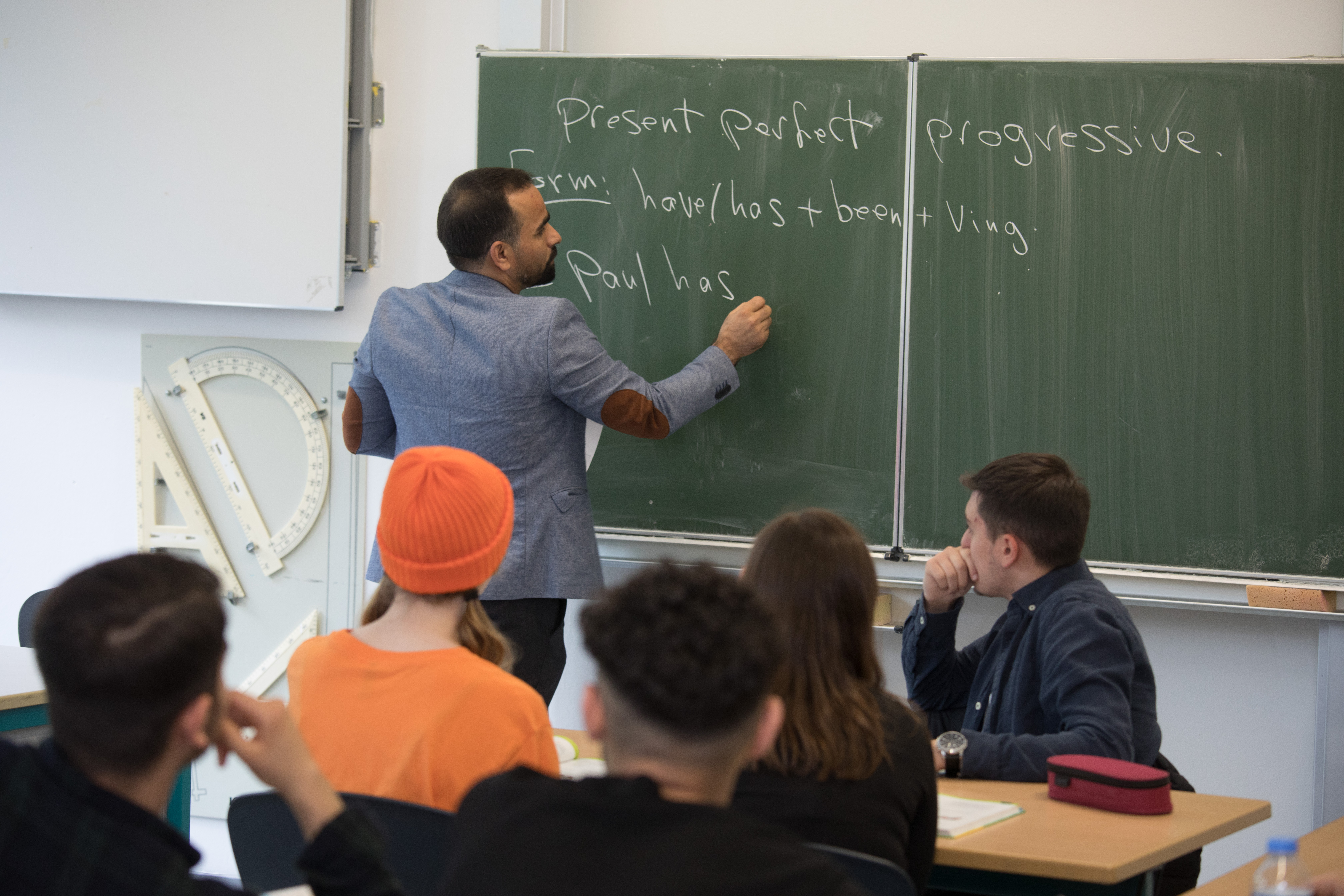 Lehrer schreibt den englischen Beispielsatz "Paus has..." mit Kreide an die Tafel. Im Vordergrund sitzen 5 Schülerinnen und Schüler, deren Blicke auf die Tafel und Lehrer gerichtet sind.